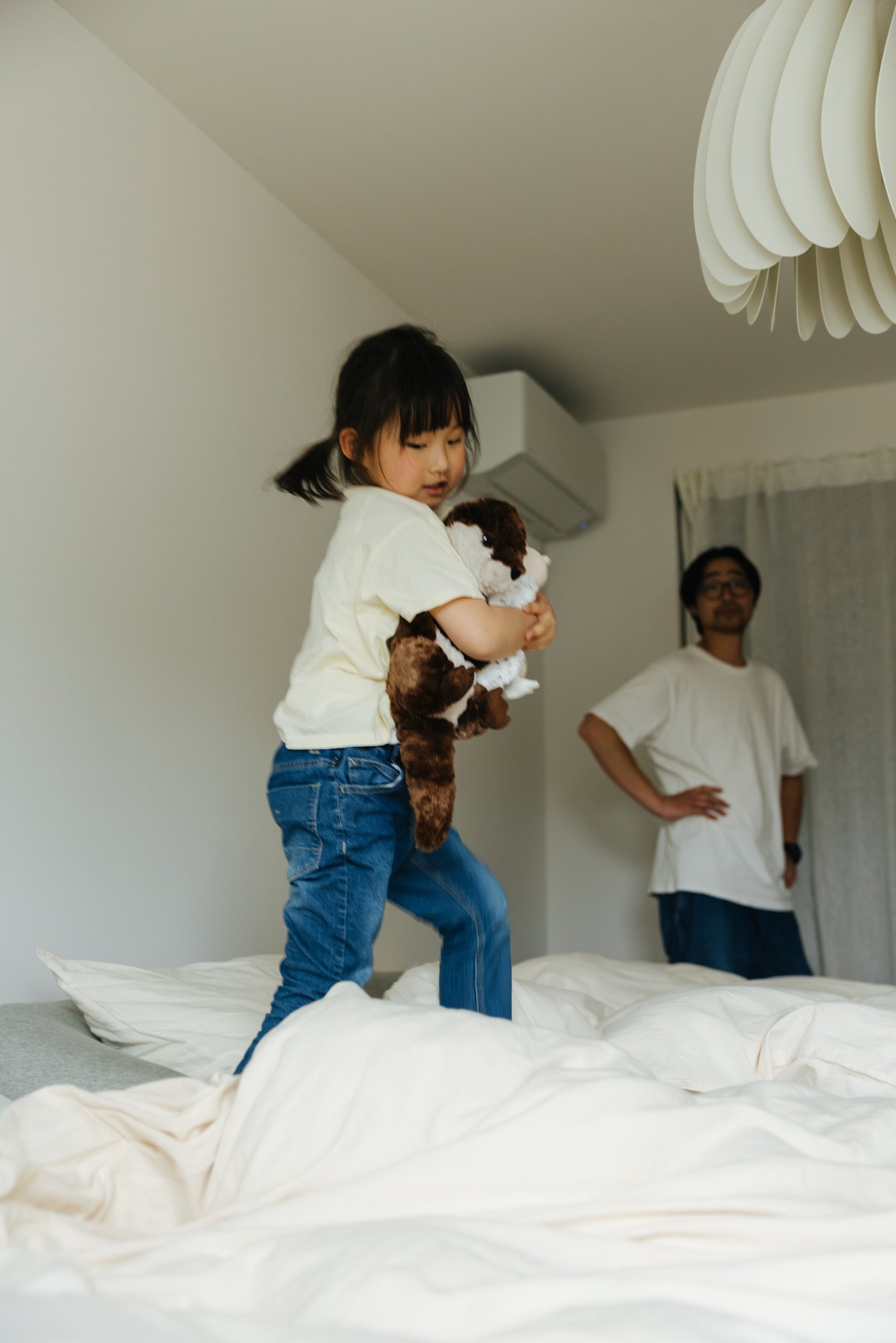 A child plays on a bed with white bed linen with a person standing behind in a white t-shirt.