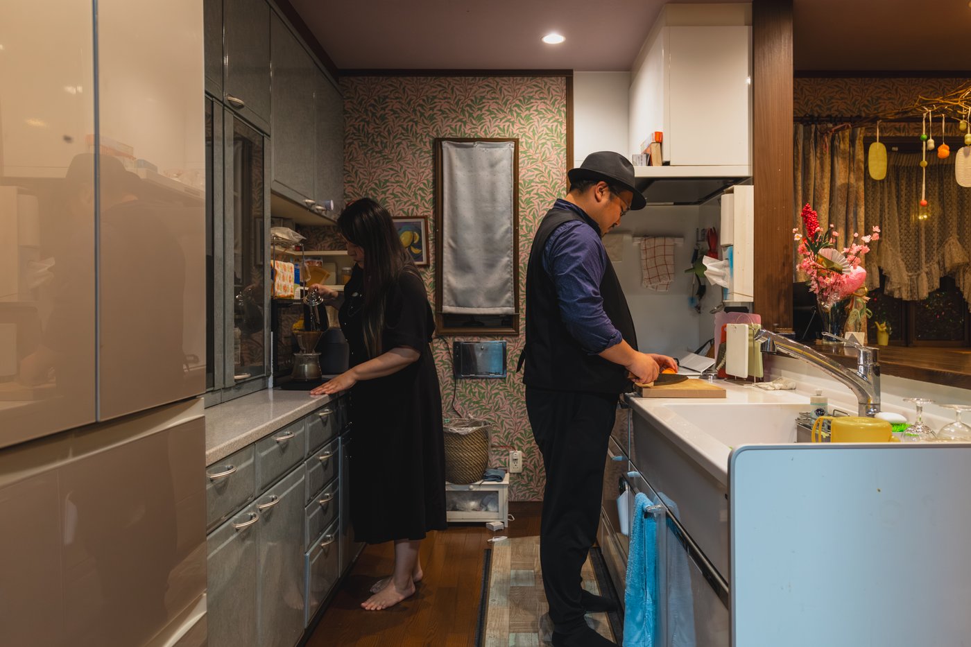 Two people cooking and preparing a drink in a grey kitchen with botanical-themed wallpaper. 