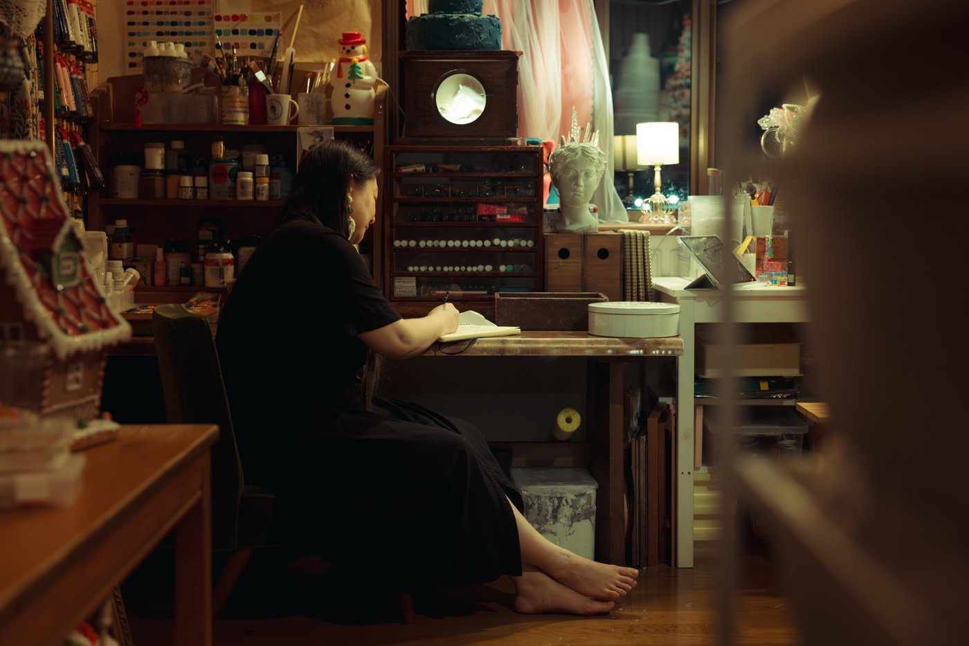 A person, seated at a desk and writing in a notebook, in a dimly lit room filled with art, ornaments, stationery and art supplies.