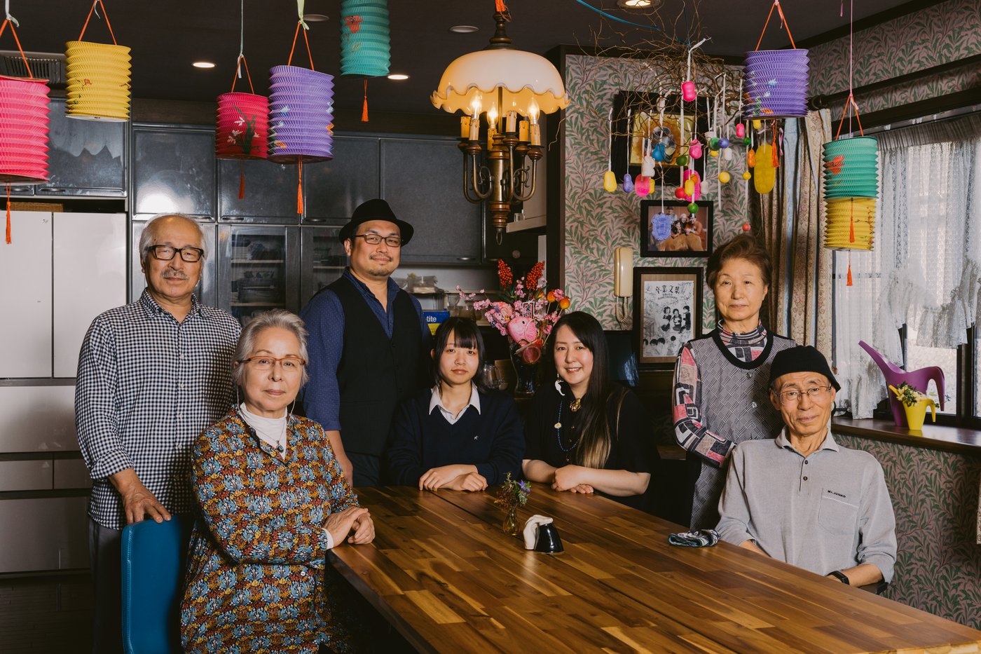 A multi-generational family sit and stand beside a wooden table in a dark kitchen with colourful paper lanterns hanging overhead.