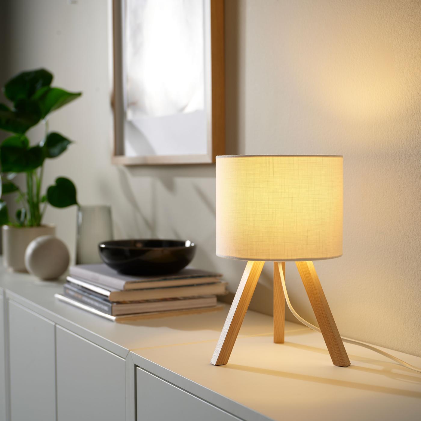 A cylinder-shaped textile table lamp with three wooden legs, emitting warm yellow light, set against a backdrop with books, plants, and framed art.