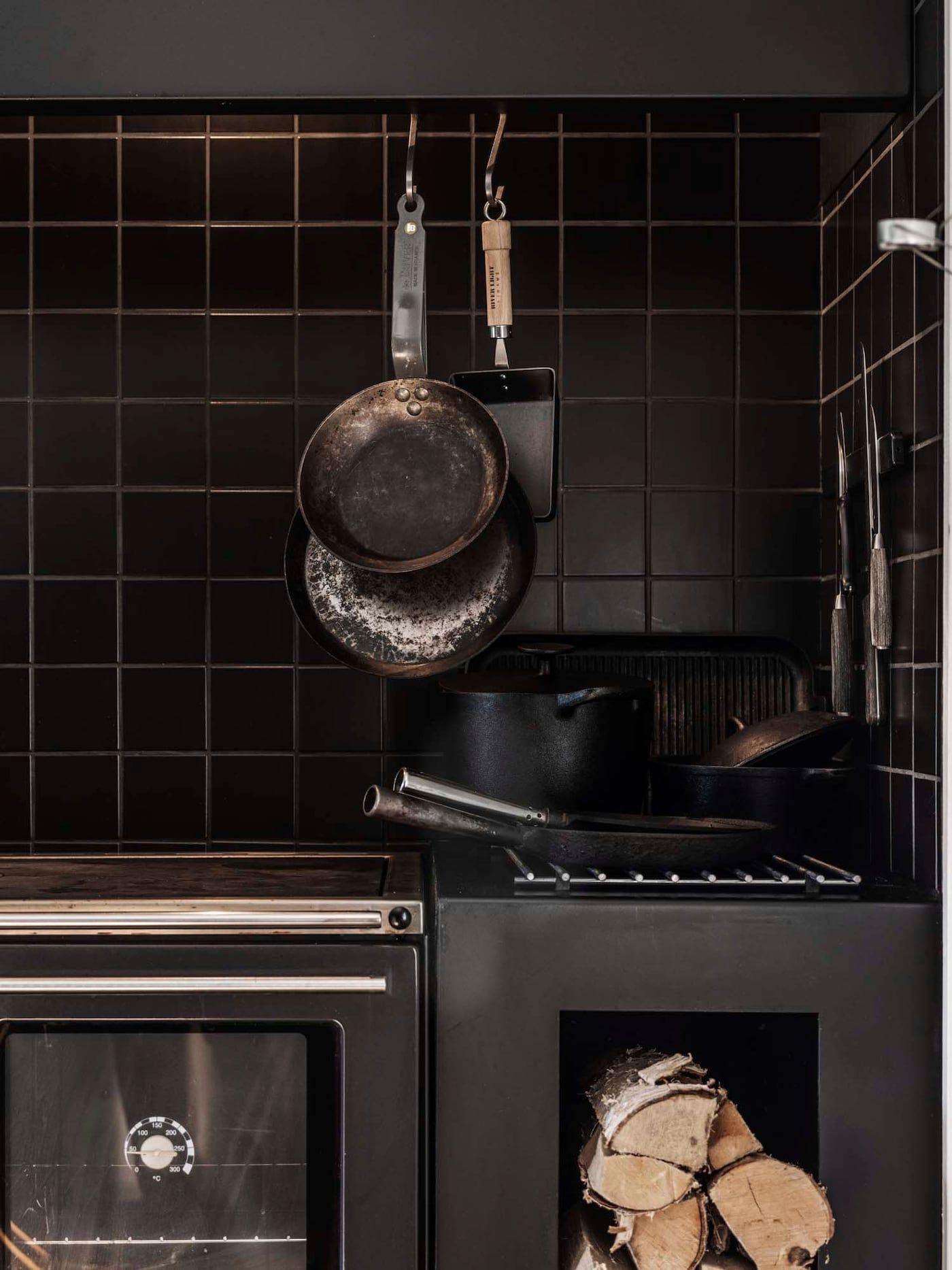 A wood-fired stove with black-tiled backsplash and cast iron pans hanging from hooks.