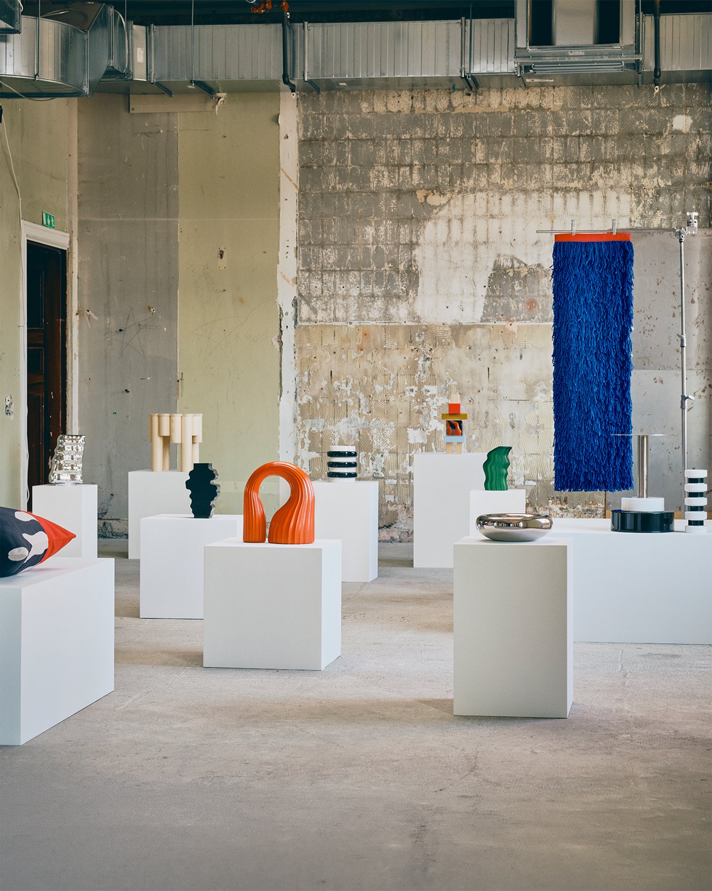 Colourful art objects are presented on white plinths in an industrial gallery setting.