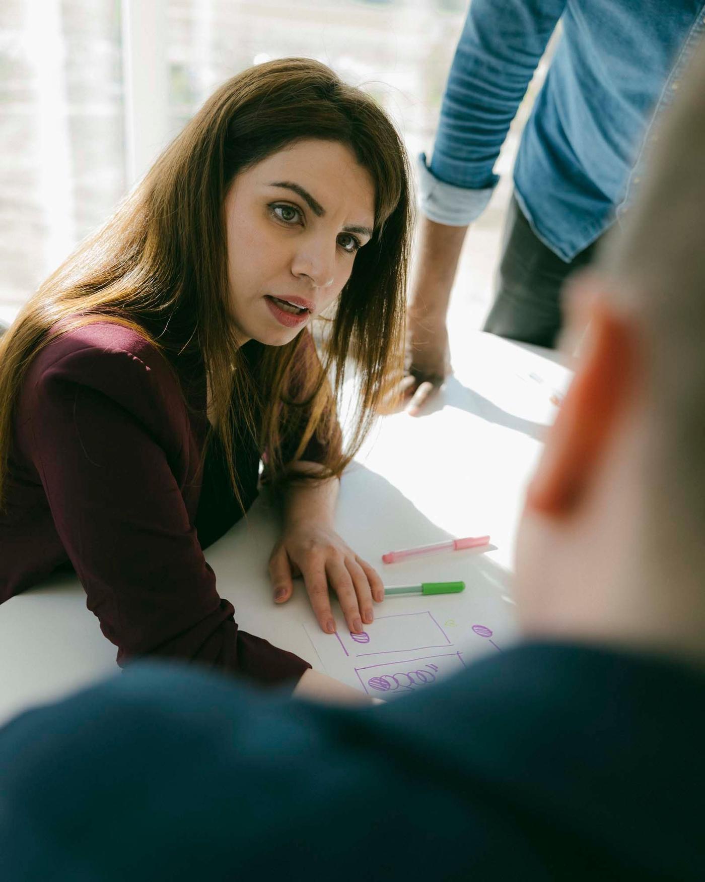 A woman sits in a meeting room. She is talking to two people. One has his back to the camera and the other person is cropped.