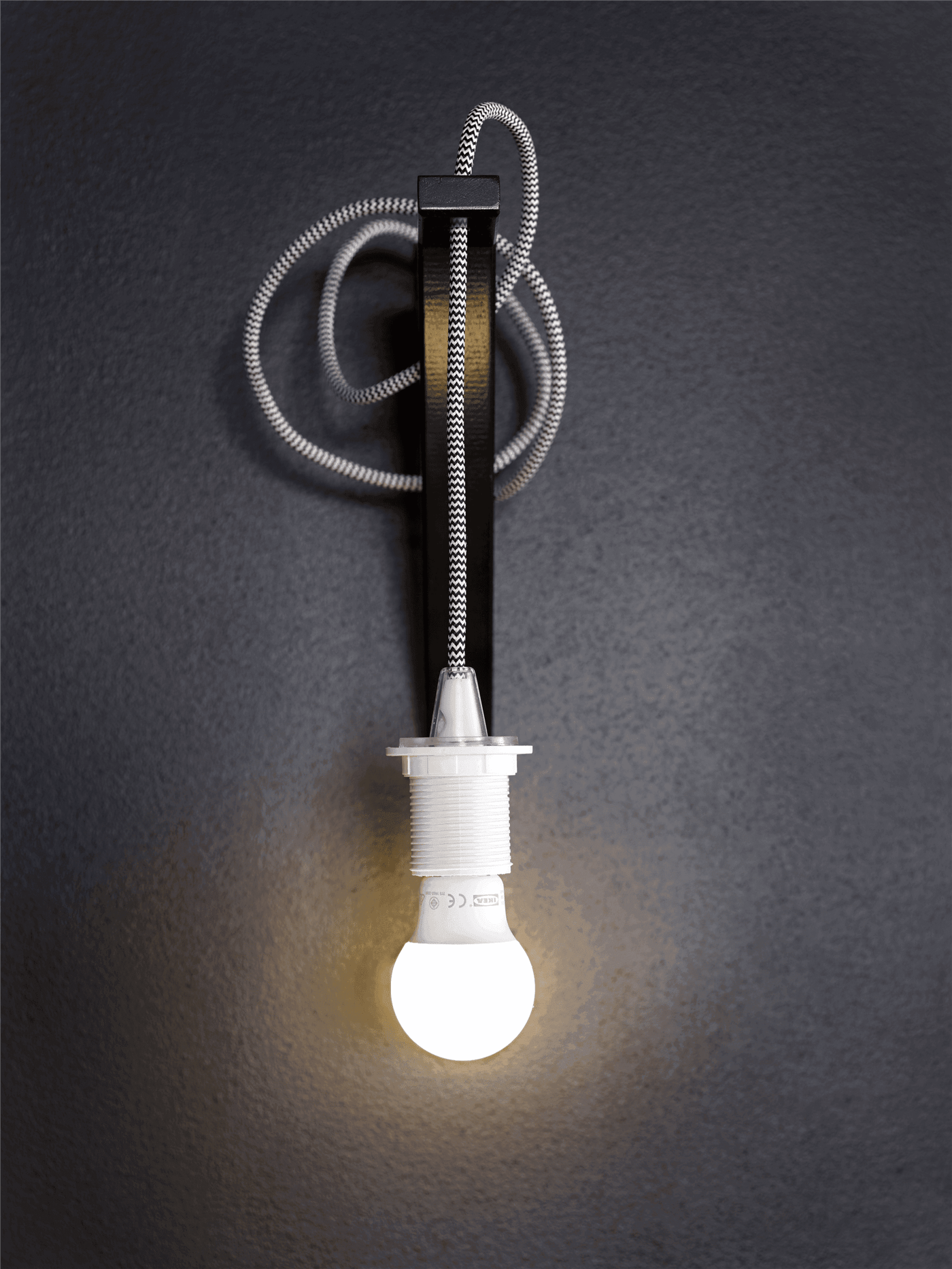 A LED lightbulb hanging in a black-and-white textile cord from a black bracket on a black wall.
