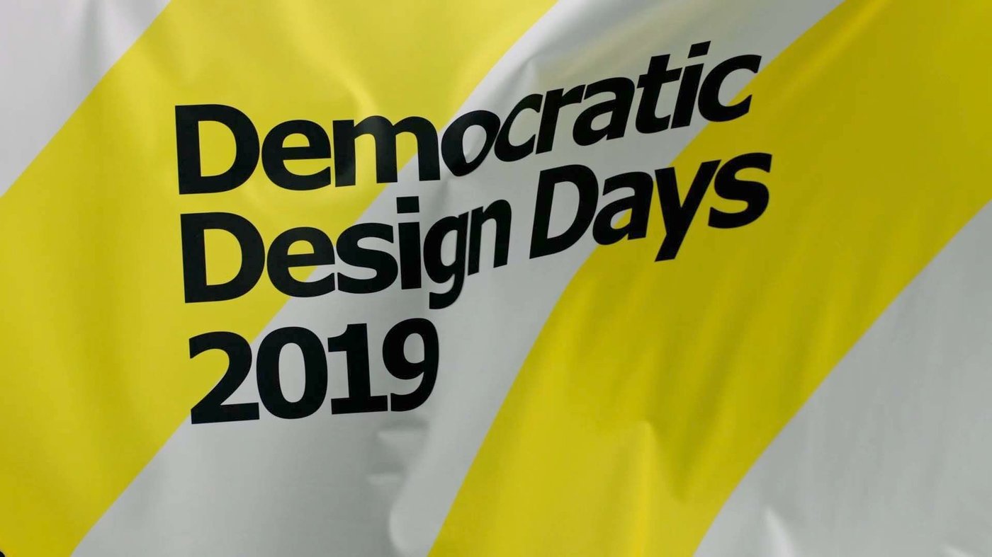 Two men sit on stools and talk to the camera.