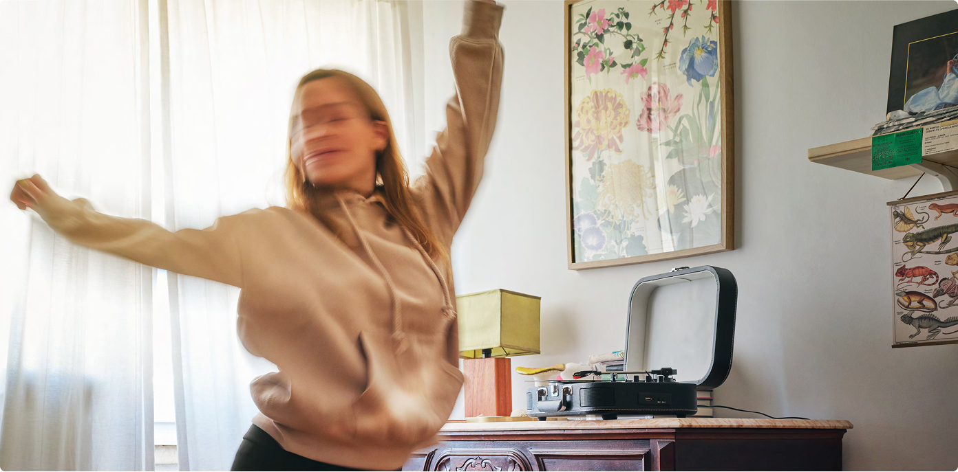 A blurred person wearing a beige hoodie, dancing beside an old-fashioned record player that's resting on a sidetable.