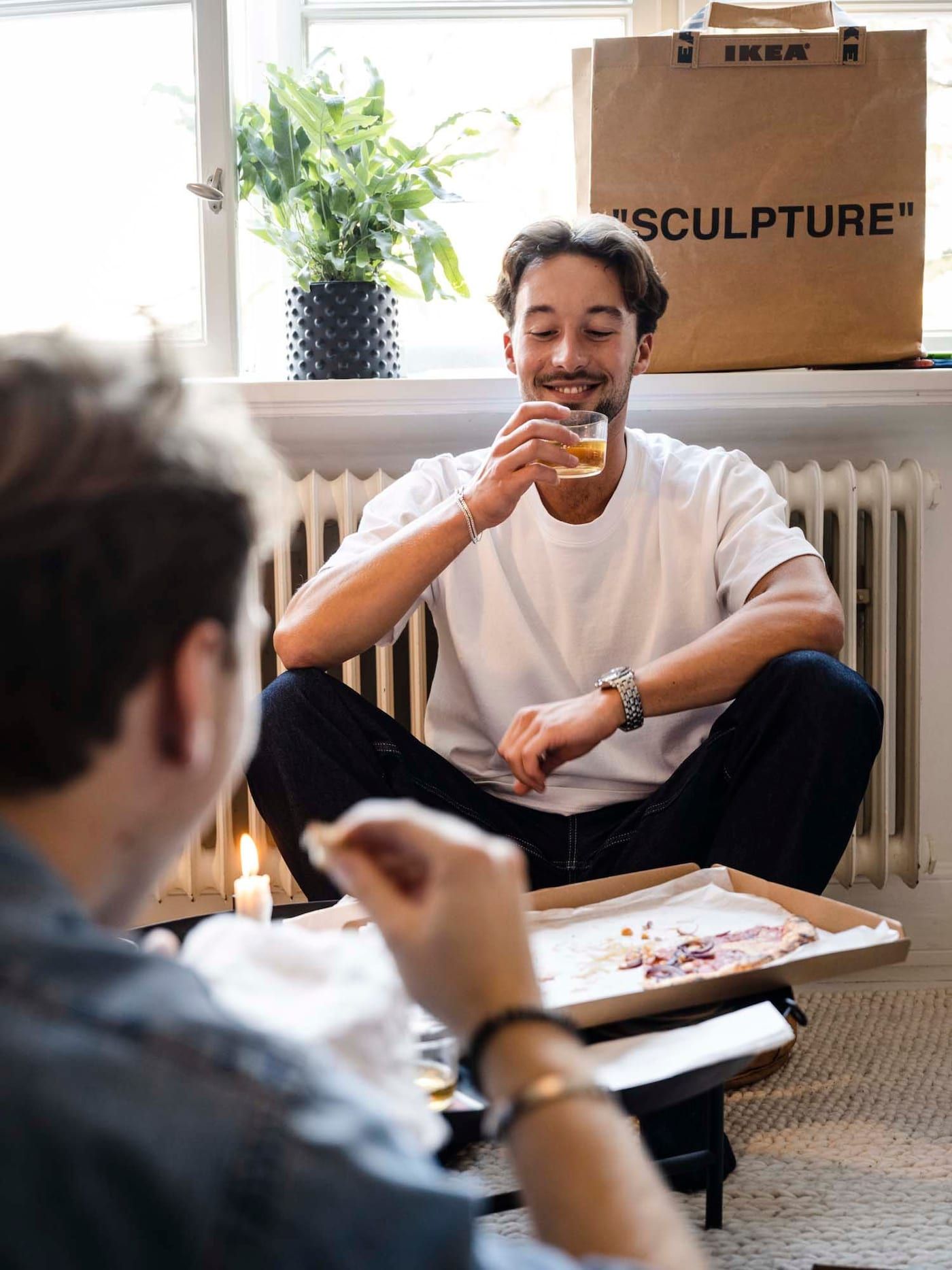 A person sits on the floor smiling while drinking from a glass, with an open pizza box at their feet. 