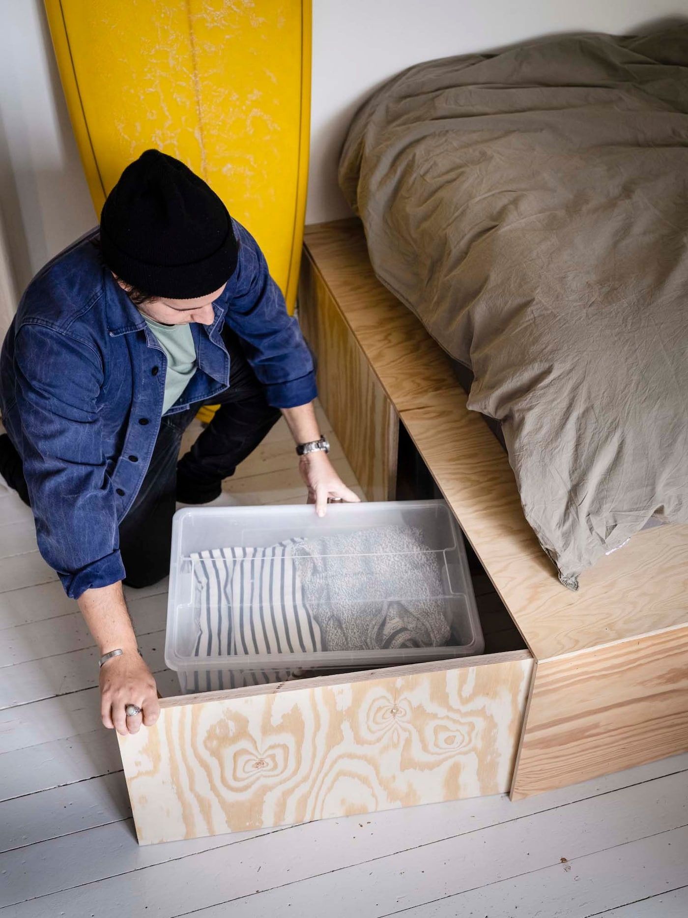 Linus opens a door in his bed's plywood base to remove a storage box containing clothes.