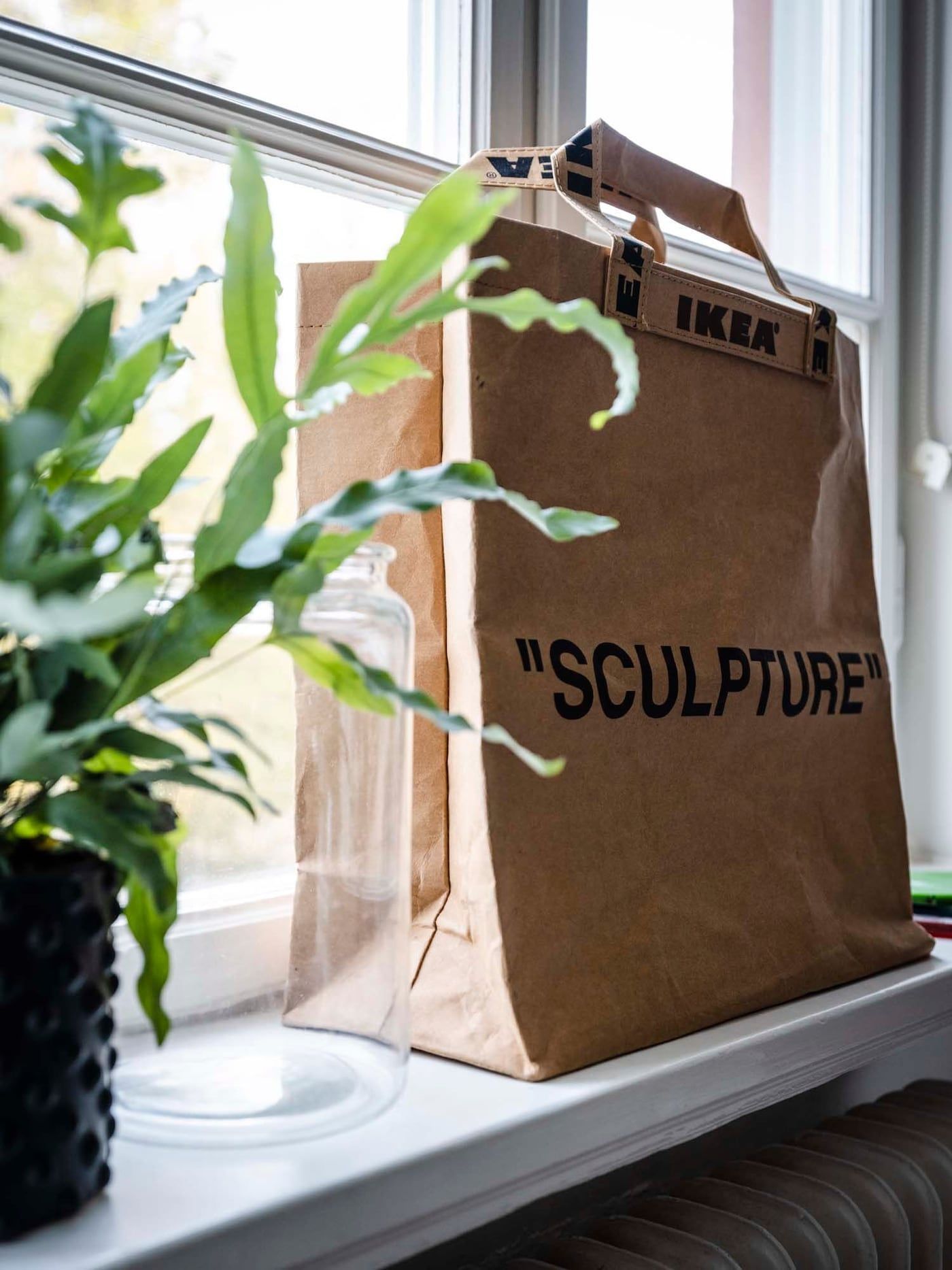 An IKEA paper bag with the word "SCULPTURE" printed on the front, placed on a windowsill.