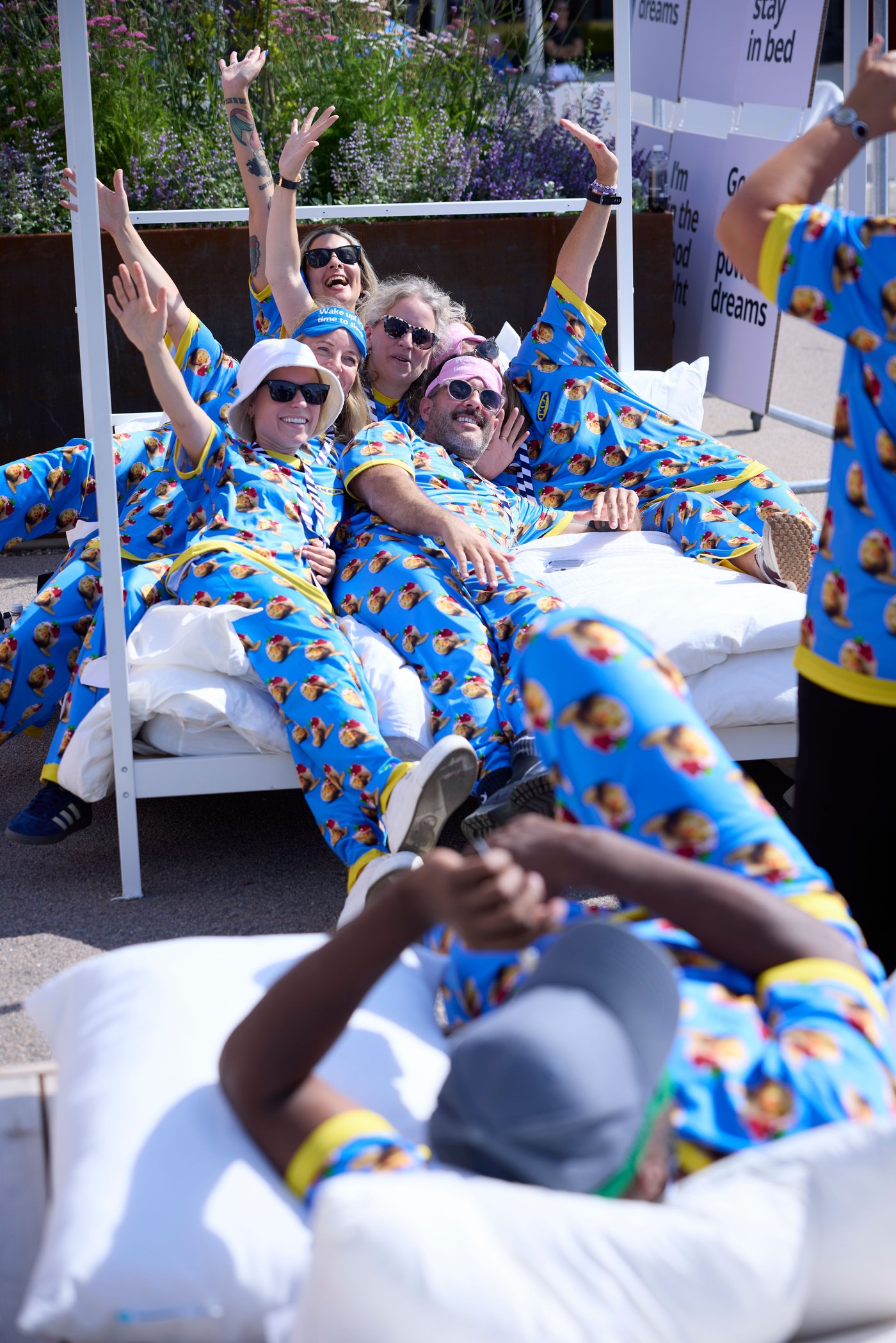 A group of people wearing identical blue pyjamas are lying in bed next to each other with raised hands and happy faces