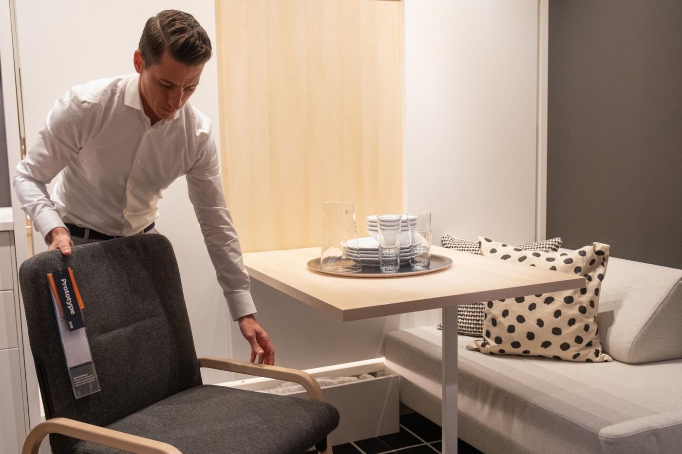 A man standing next to a table and an armchair.