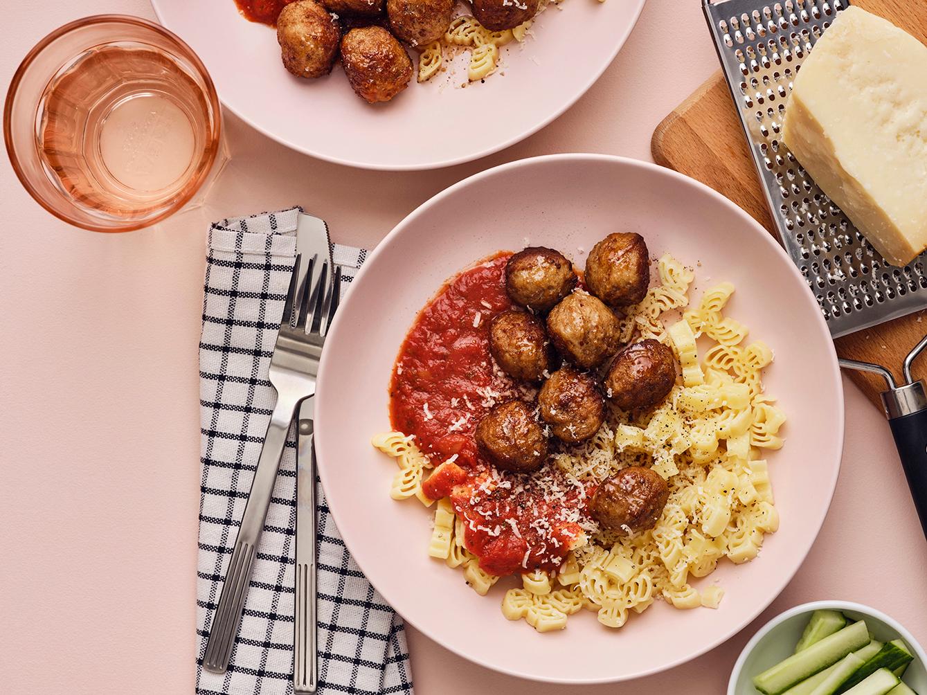 IKEA meatballs served with pasta sauce and pasta on two pink plates on a pink table.