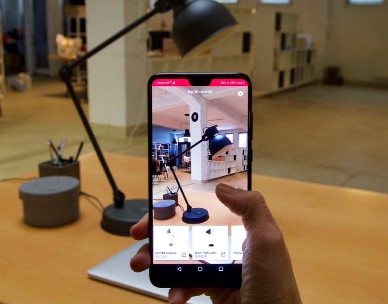A hand holding a phone with the Nyris app takes a photo of a black desk lamp.