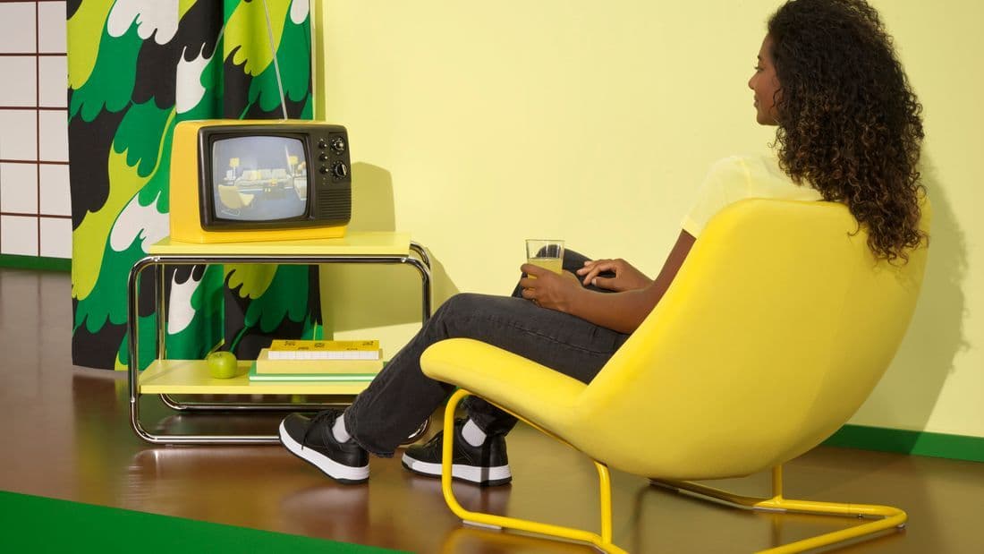 A person sits in a yellow lounge chair in front of a vintage television. Behind the television you see curtains with a bold green, black and white pattern.