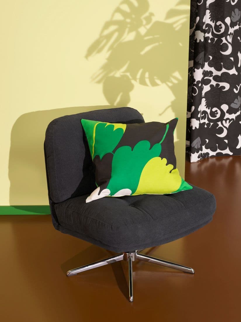 A picture of a black lounge chair, on it a pillowcase with pattern. Behind the chair there is a curtain with pattern. All products are part of the Nytillverkad collection.