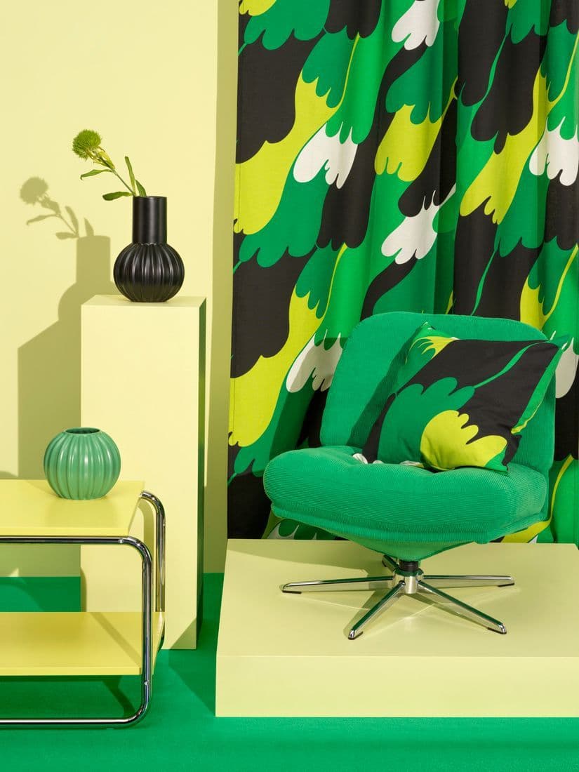 This picture showcases a room set up of the Nytillverkad collection consisting of a curtain with pattern, a green lounge chair with a pillow on it that has the same pattern as the curtain.