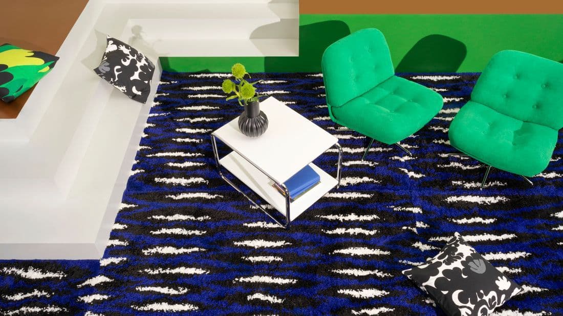 A roomset showing the Nytillverkad collection consisting of a blue-black-white rug, two green lounge chairs and several pillow with different patterns.
