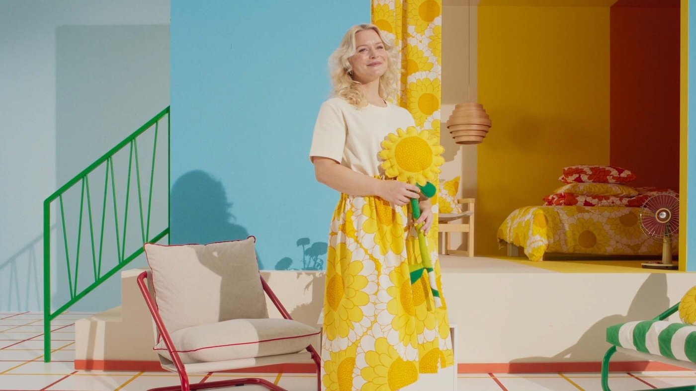 A still of a video. A woman holds a fabric sunflower in a roomset. She is smiling.