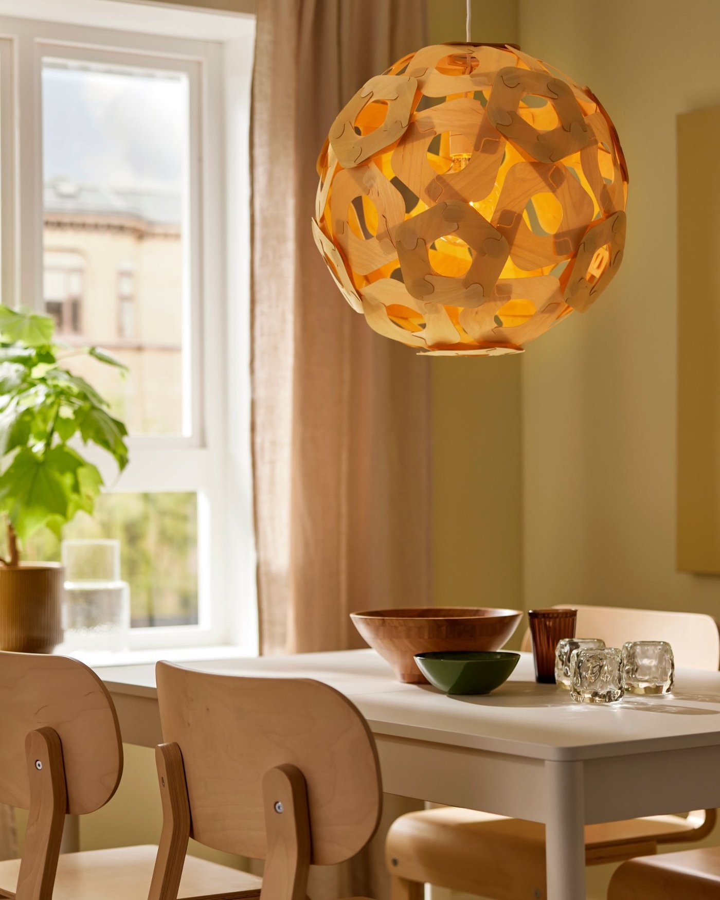 A spherical wooden look pendant lamp hanging from a ceiling, glowing near a window with earthy walls and plant beside.