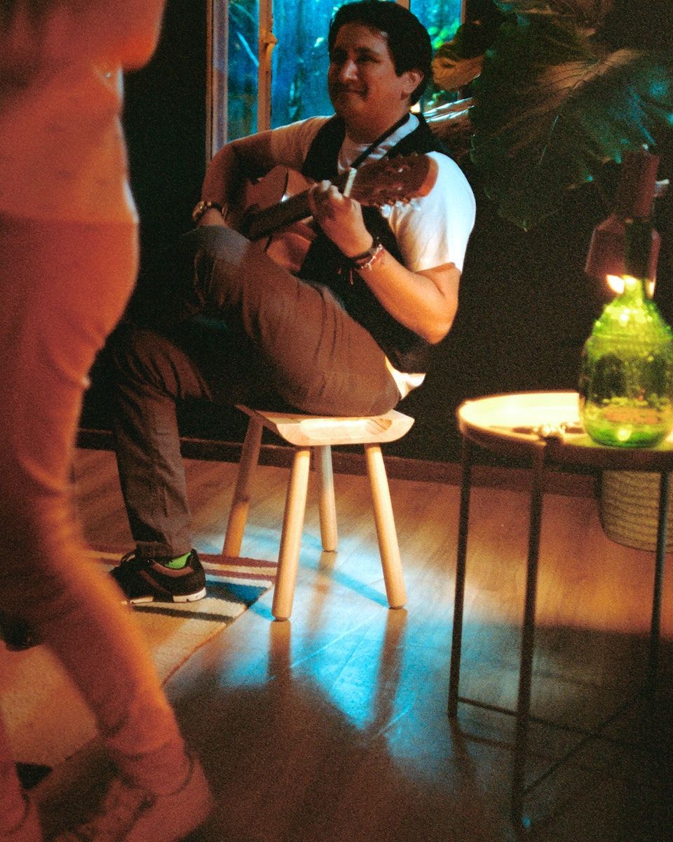 A person seated on an ÖMSESIDIG stool plays the guitar in a dark room of dancing people.