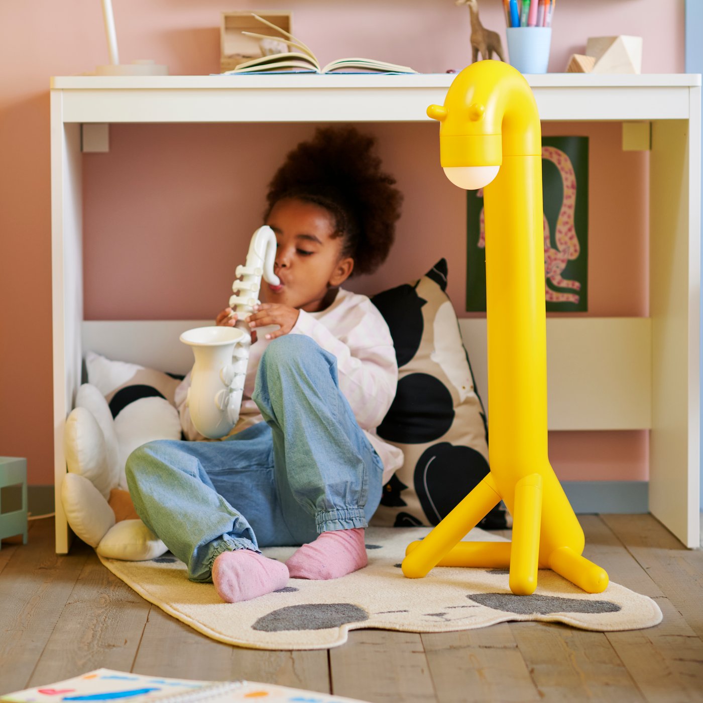 A child sitting underneath a desk on top of a rug and and some cushions while they play a white saxophone.