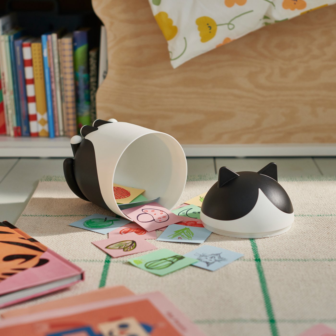 The GREJSIMJOS cat storage container in black and white, laying on the floor with coloured paper pieces spilling out and around it.