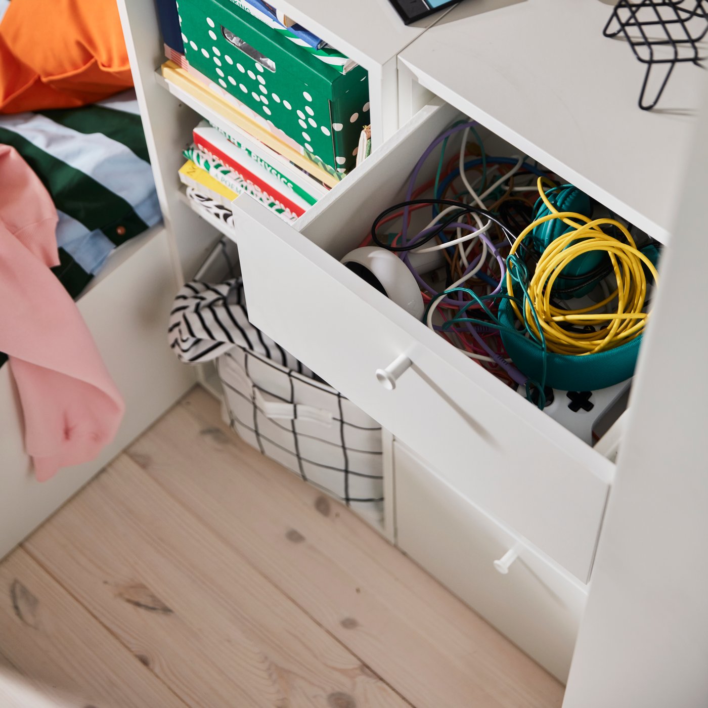 A small chest of drawers in white with open storage filled with books and boxes, as well as an open drawer full of rolled up cords and accessories.