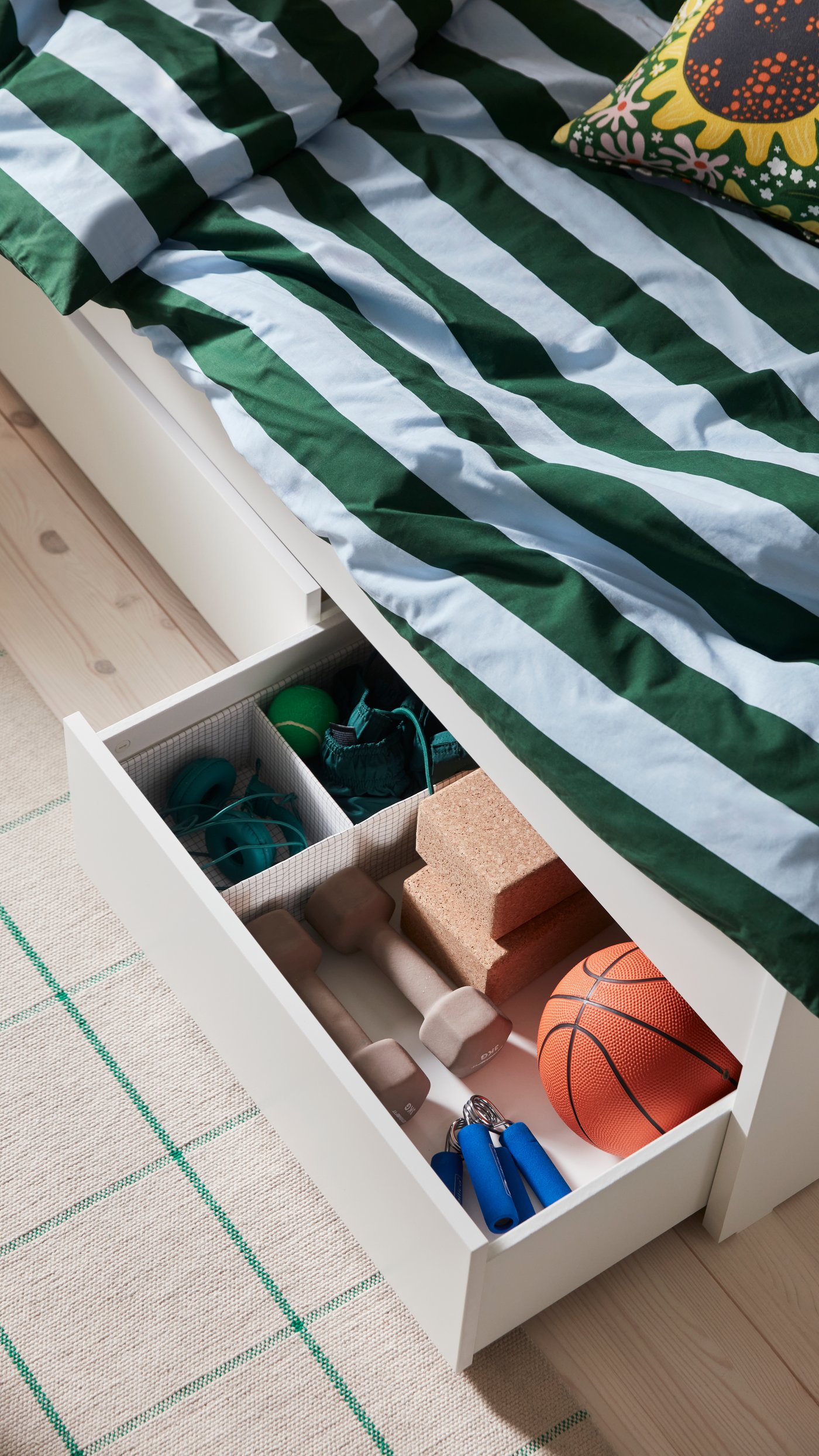 A bed with storage and green and white striped bedding, with an open drawer that holds yoga accessories in smaller organised boxes.