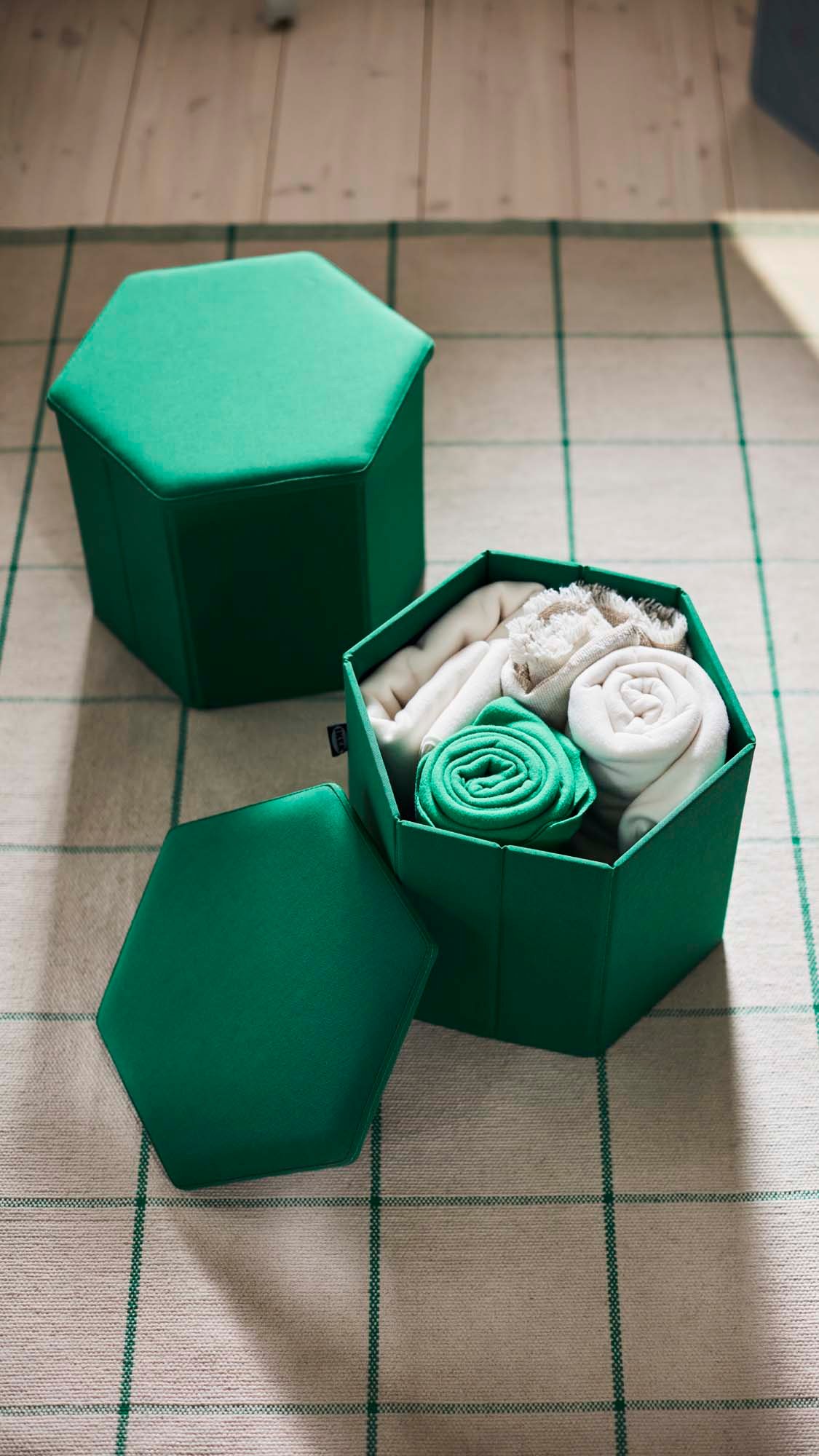 Two green KJUGE pouffes, one closed, one with the lid open full of rolled towels, sitting on a cream and green striped rug.