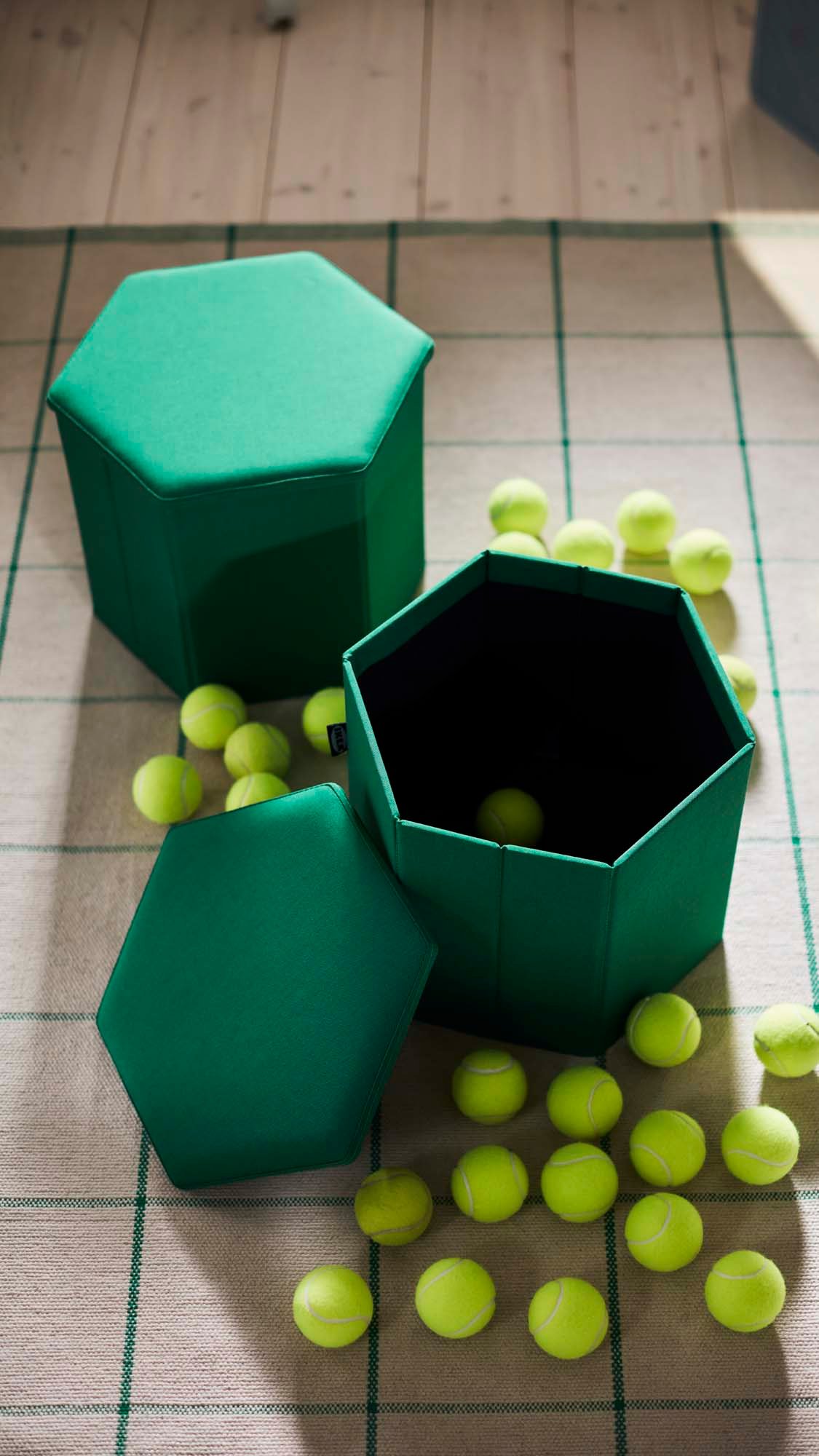 Two green KJUGE pouffes, one closed, one with the lid open and tennis balls in and around it, sitting on a cream and green striped rug.