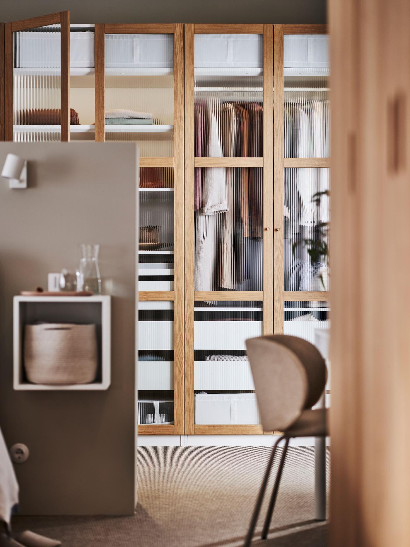 An open door reveals a PAX wardrobe system in a bedroom. The wardrobe has wood framed glass doors. In the wardrobe are drawers and clothes hanging from rails.