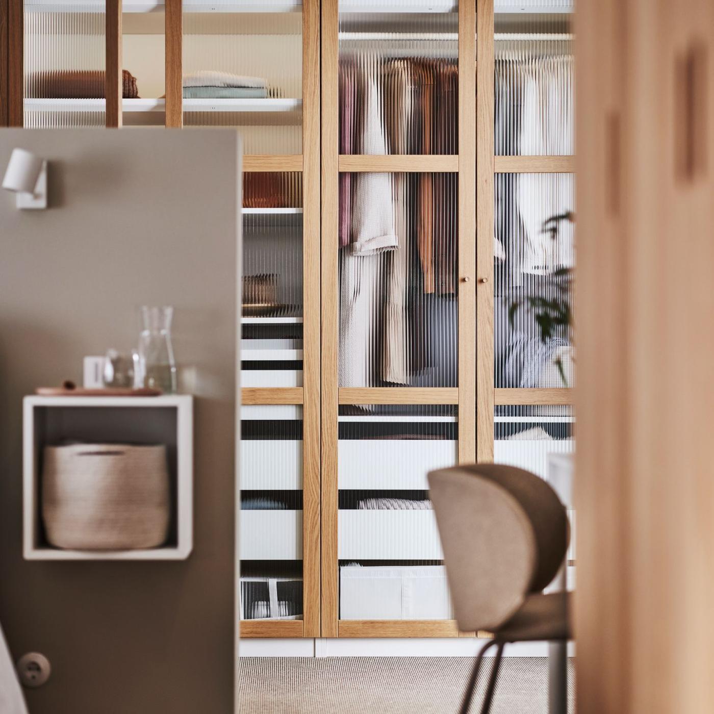 An open door reveals a PAX wardrobe system in a bedroom. The wardrobe has wood framed glass doors. In the wardrobe are drawers and clothes hanging from rails.
