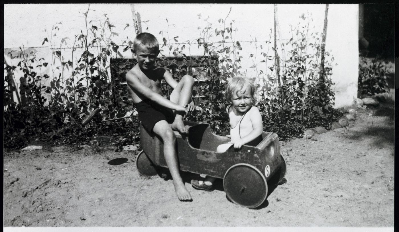 A childhood black and white photo of Ingvar Kamprad and his sister outdoors. Ingvar sits on a bench and his sister sits in a wooden car.