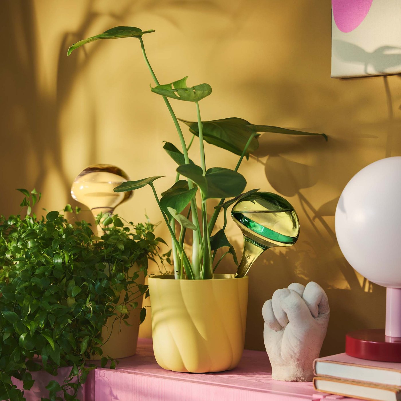 A yellow plant pot on a pink unit, in fronto f a yellow wall. In the plant pot is a green plant and a DOFTRIPS self-watering insert.
