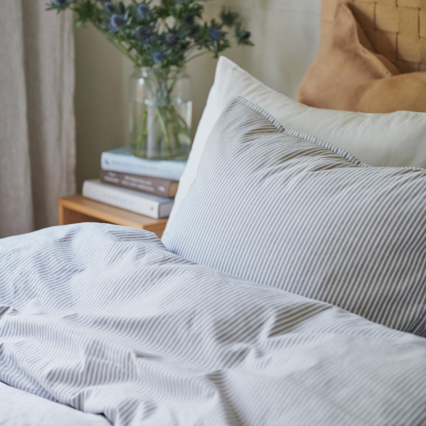 Close up of SOLFIBBLA bed textiles in a white and blue stripe, layered on a white and tan pillow and woven headboard.