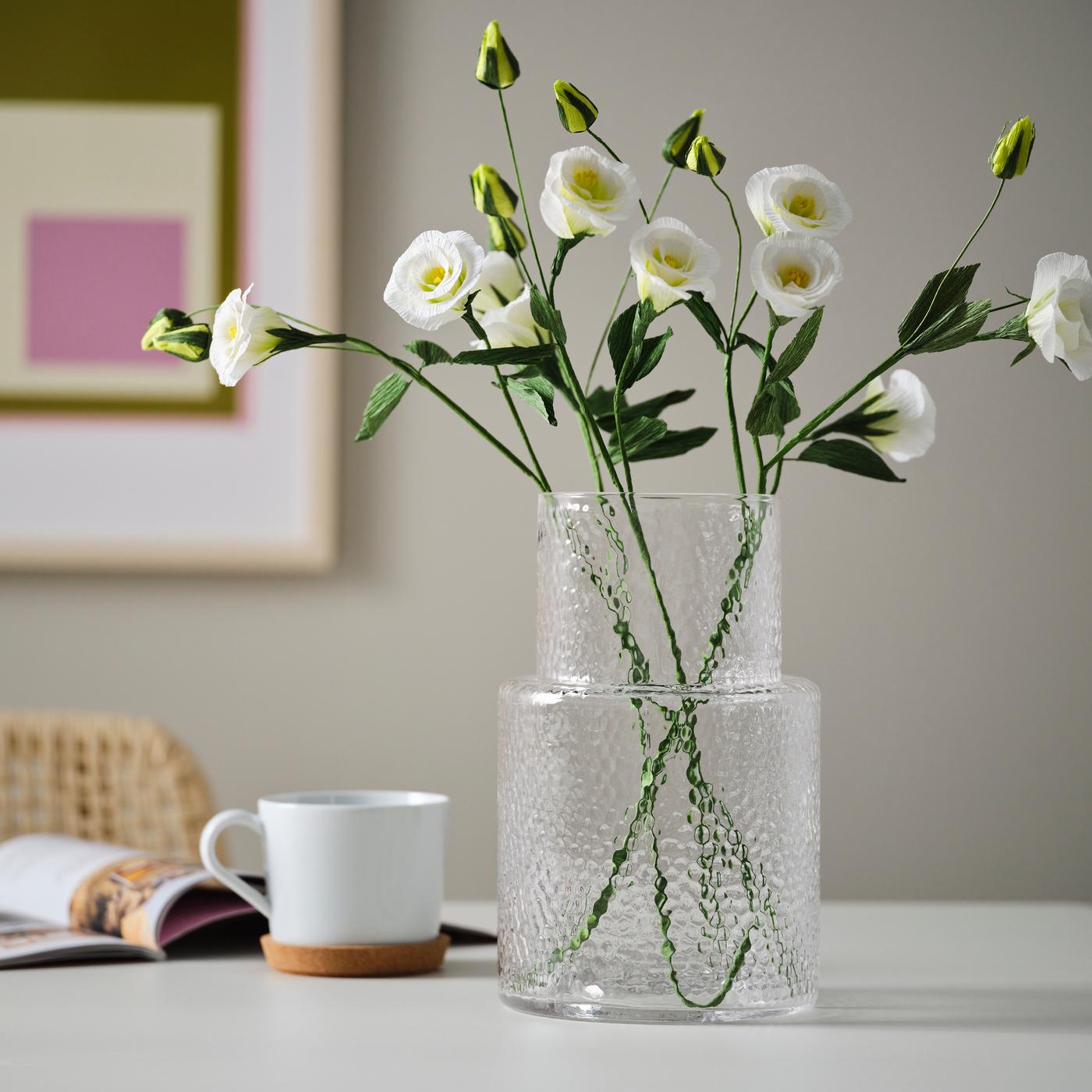 A dining room with a clear vase filled with white GULDSPETT artificial flowers, placed on a table with a painting in the background.