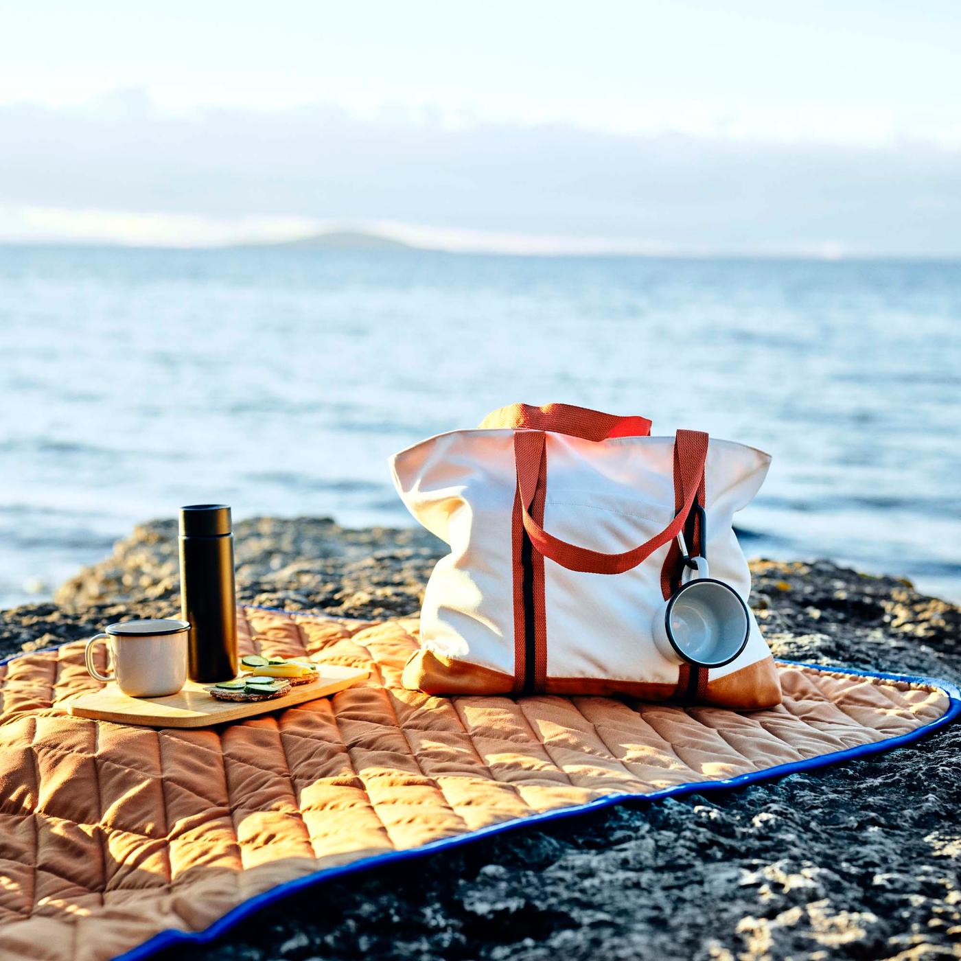 A white canvas bag with orange handles on an orange quilted picnic blanket on a cliff by the sea. 