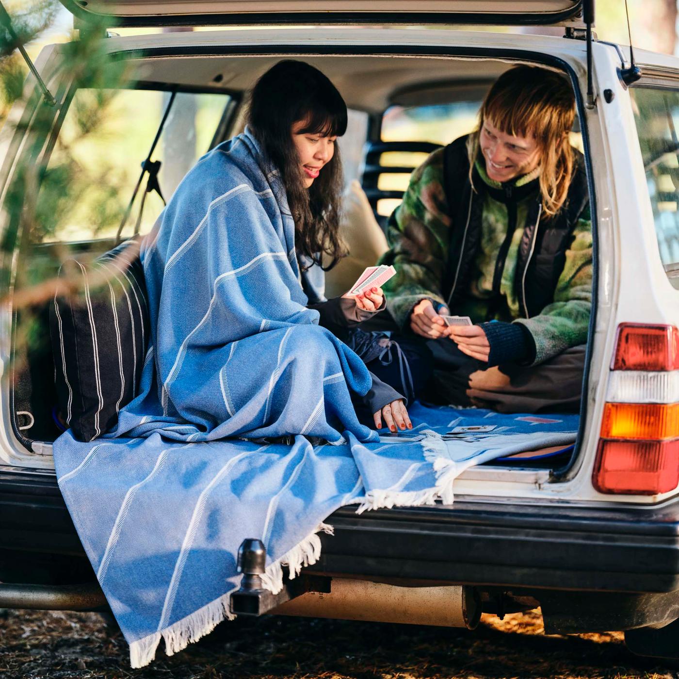 Two people play cards in an open boot of a car, one is wrapped in a blue and white throw.