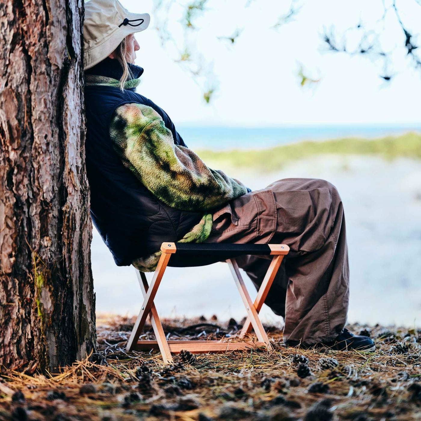 A person sitting on a stool leans against a tree trunk by the sea.
