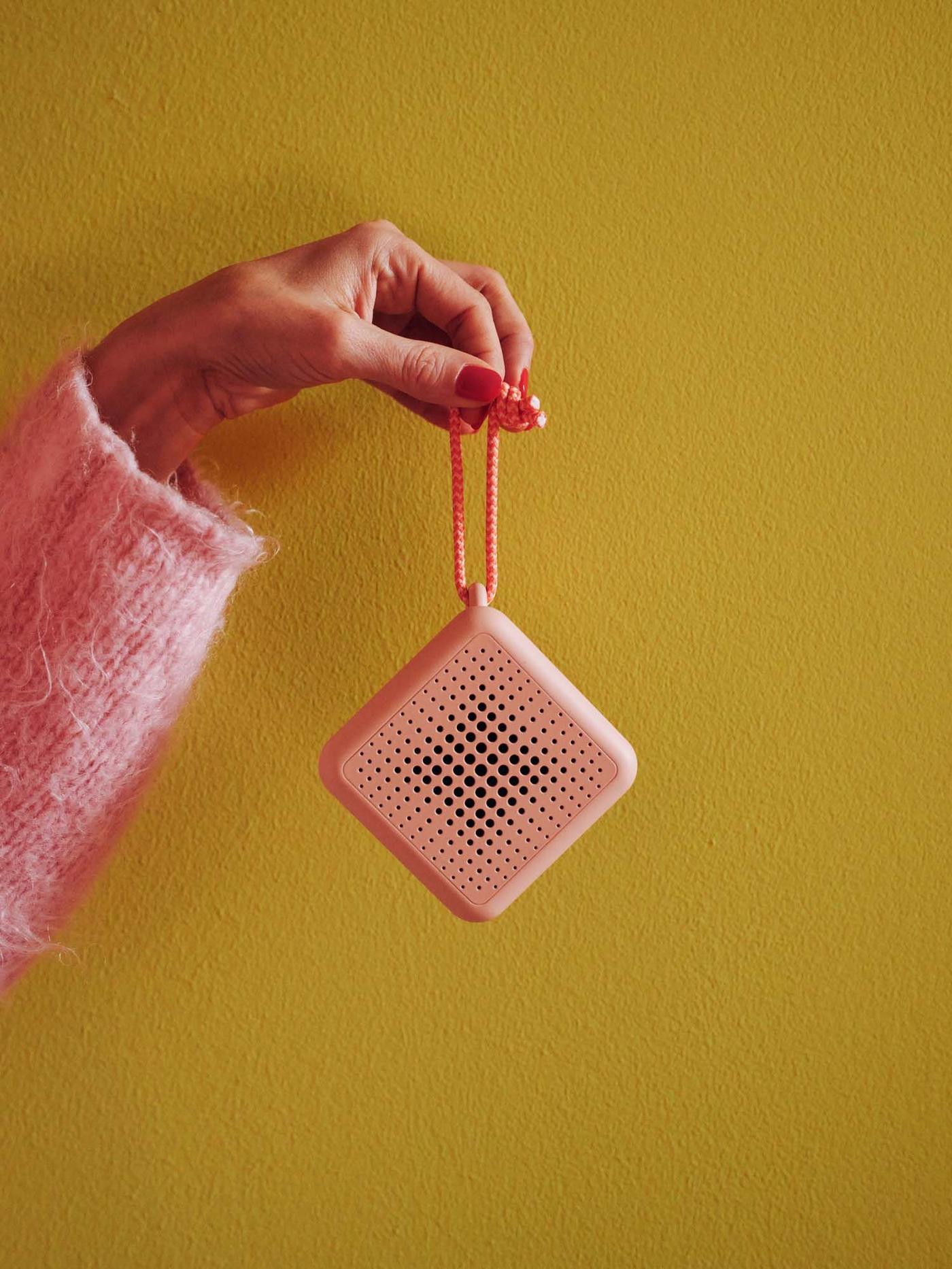A hand with a pink sleeve holds a pink VAPPEBY Bluetooth speaker against an ochre yellow background.