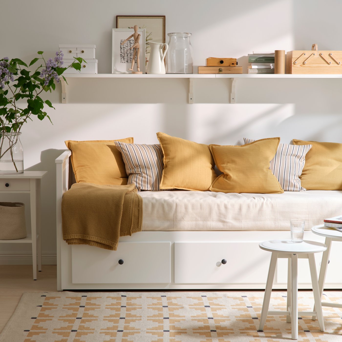 A white, bright room with a white HEMNES daybed against the wall, decorated with yellow cushions and throws. 