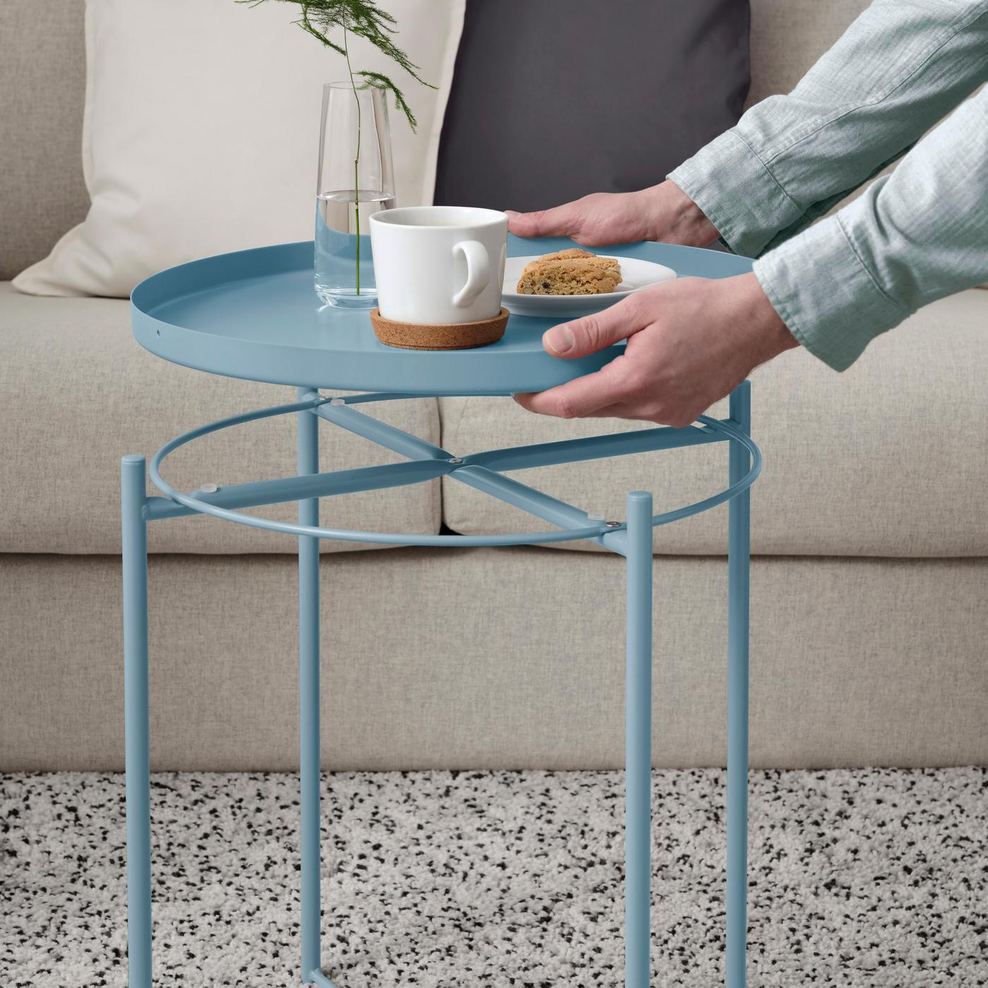 A person about to place the tray part of the GLADOM side table in light blue down, with a white coffee mug, pastry on white dish and vase with greenery.