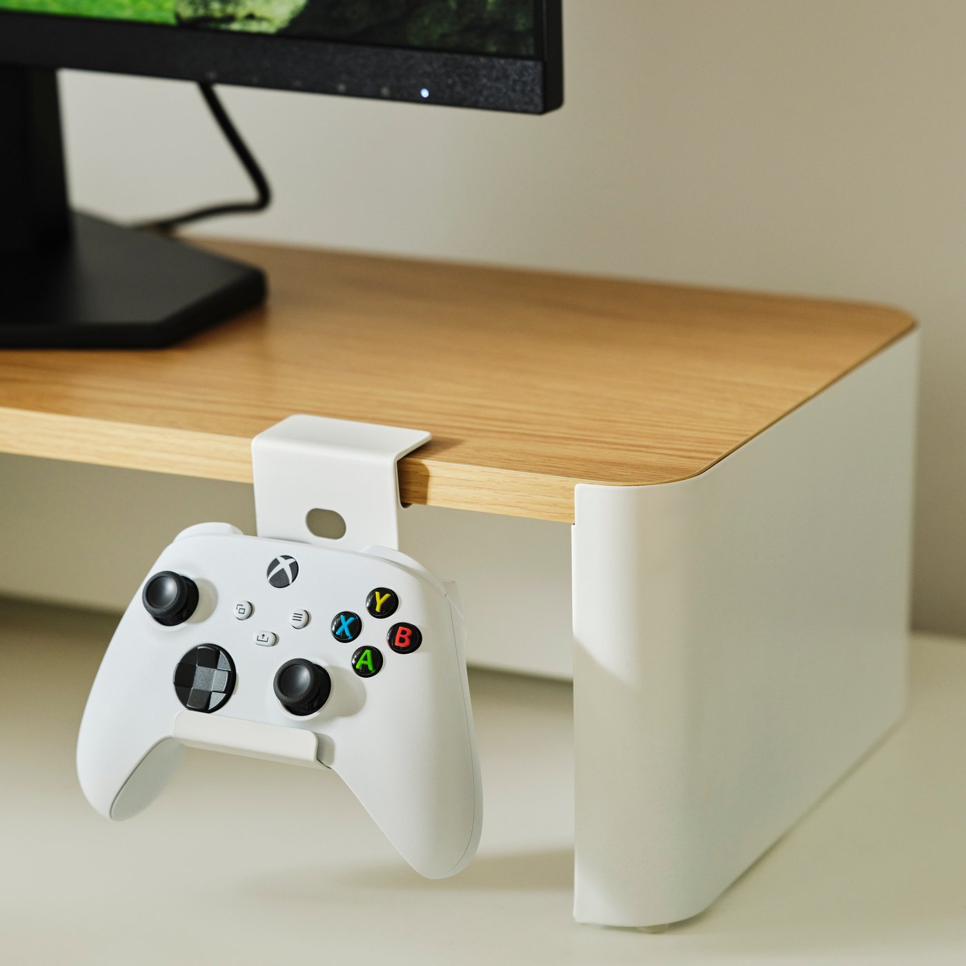 A white controller holder attached to the edge of a monitor riser, holding an Xbox controller beneath a monitor.