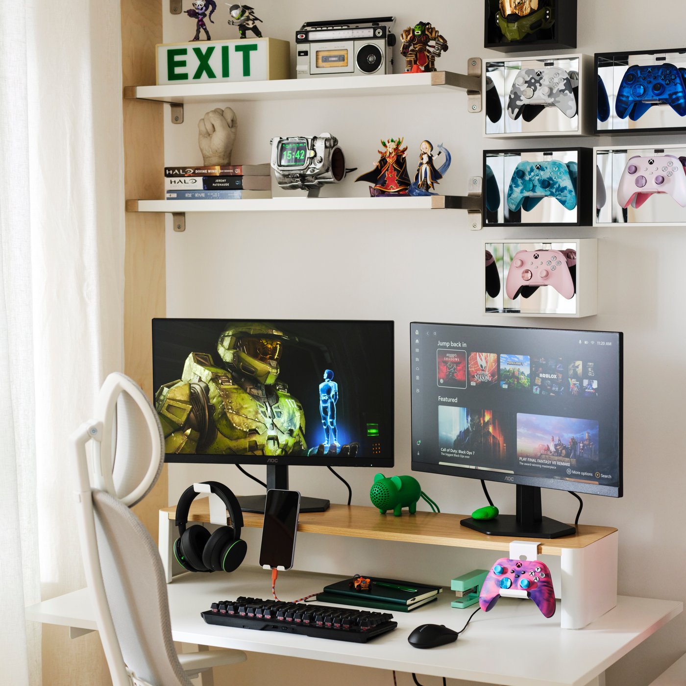 A monitor stand supporting two computer monitors above a desk, with space underneath for headphones, a phone, notebooks and gaming accessories.