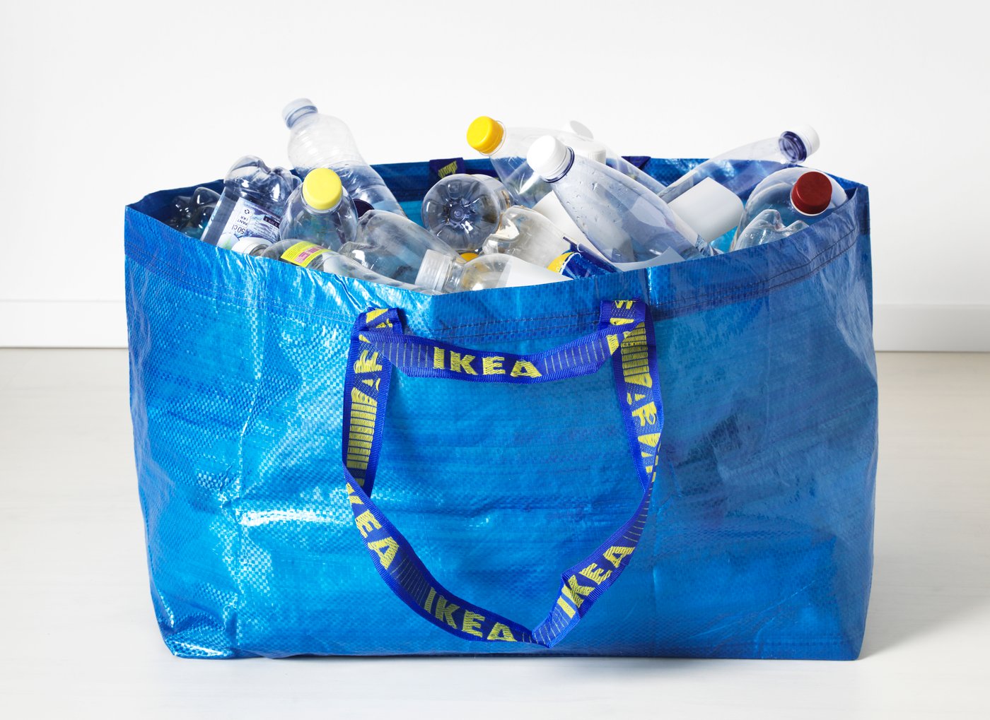 A blue FRAKTA bag filled with empty PET bottles on a floor, against a white background.