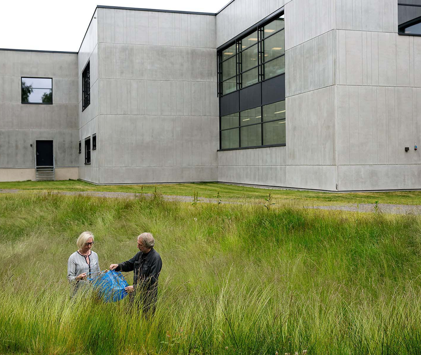 Marianne Hagberg and Knut Hagberg stand in a field of long green grass in front of a large concrete building. They are opening a FRAKTA bag.