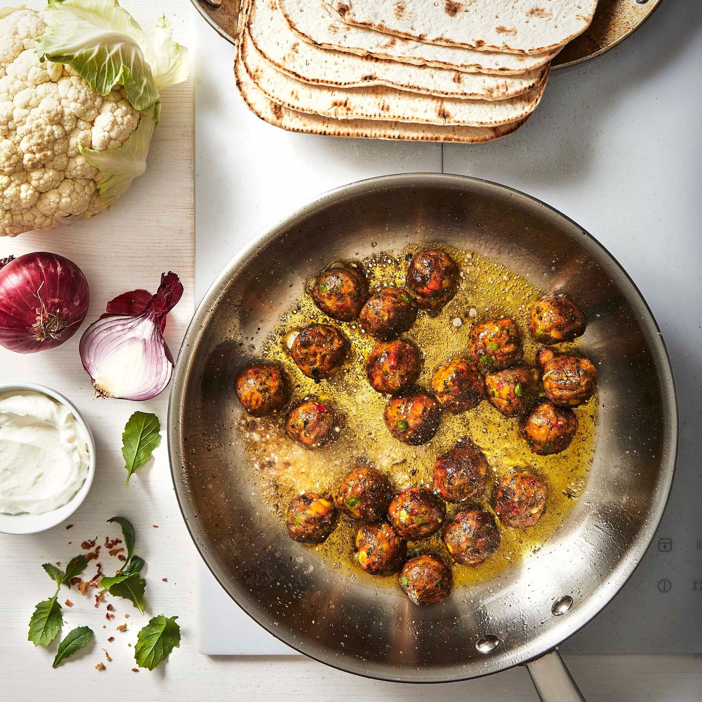 A frying pan filled with HUVUDROLL vegetable balls in a curry sauce, with vegetables, herbs and bread on the side.