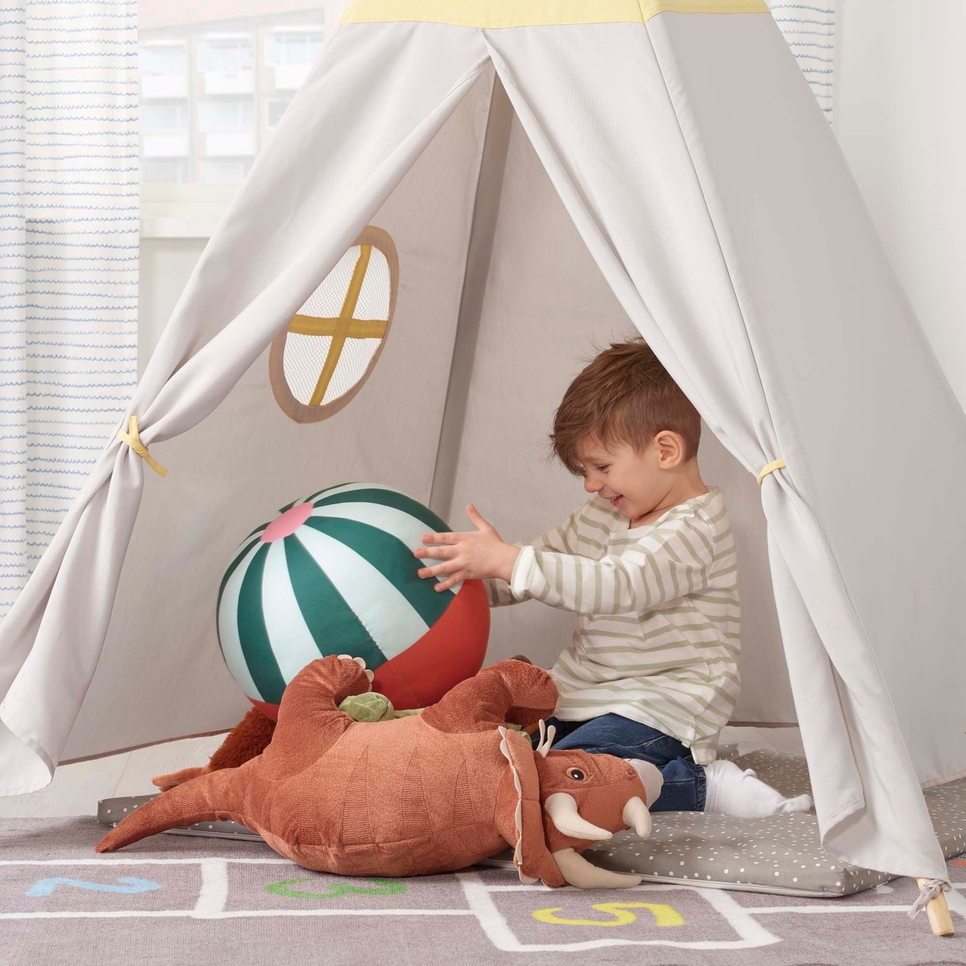 A child sits in a white teepee. He is playing with a striped ball and a dinosaur.