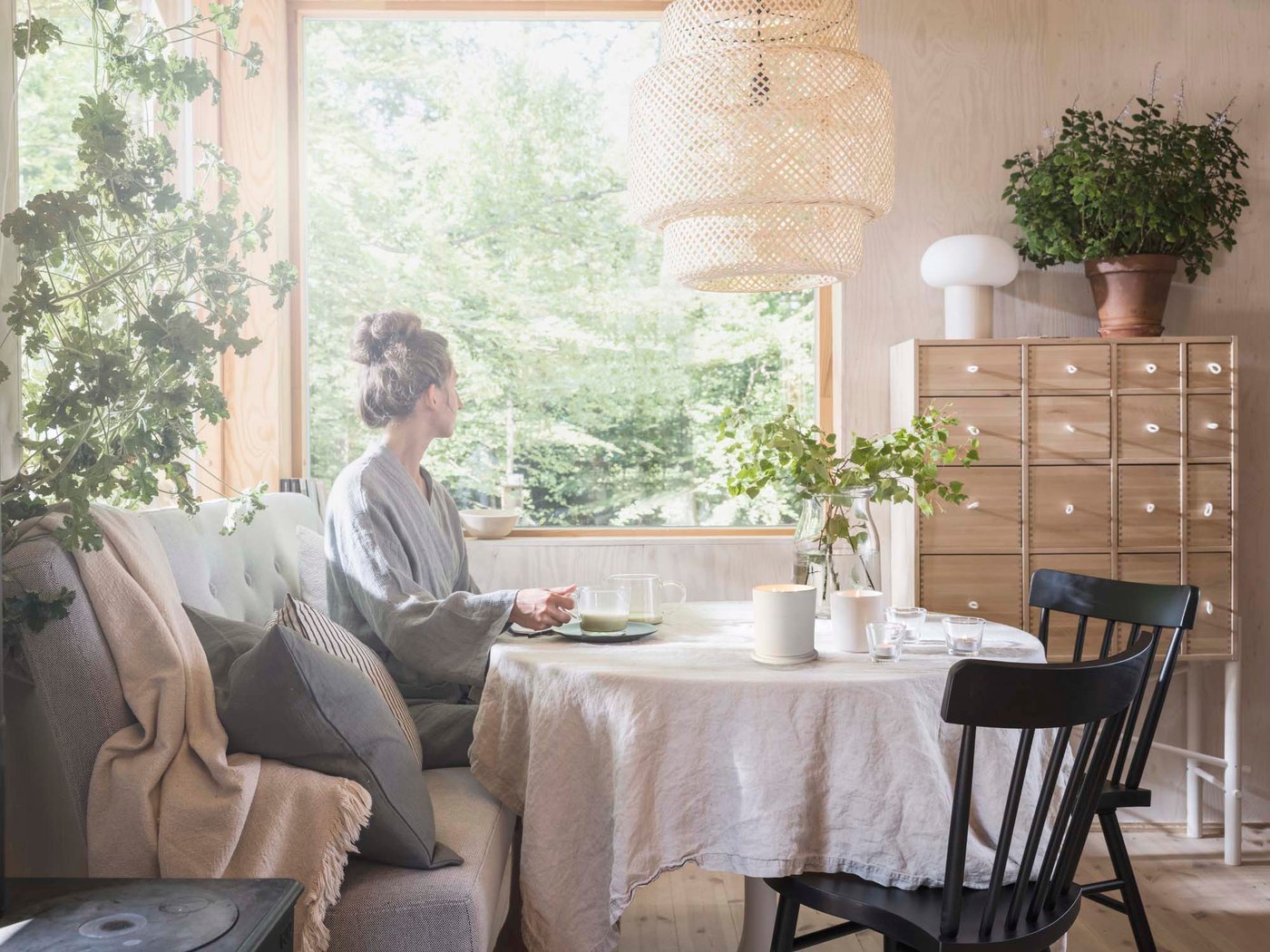 A person sits at a table in a calm room with natural materials and several plants, and looks out one of the windows.